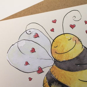 Op de afbeelding: Een aquarel schilderij van een cartoon bij met gesloten ogen en een glimlach. De bij heeft zwart-gele strepen en witte vleugels. De bij is omringd door kleine rode hartjes.