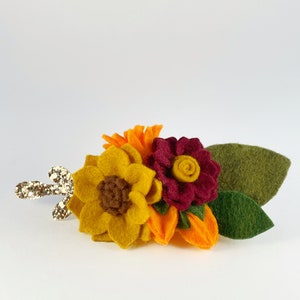 Op de afbeelding: Een haarspeld van vilt met een zonnebloem, een bordeauxrode bloem en oranje bloemen. De speld heeft een gouden glitter steel.