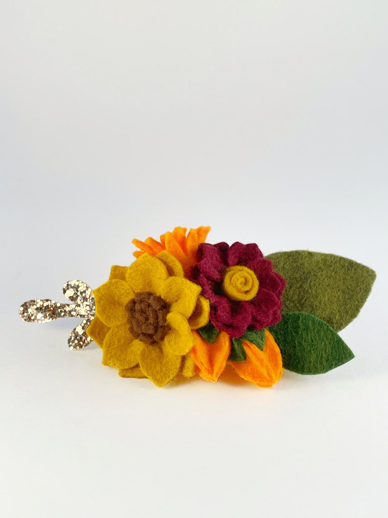 Op de afbeelding: Een haarspeld van vilt met een zonnebloem, een bordeauxrode bloem en oranje bloemen. De speld heeft een gouden glitter steel.