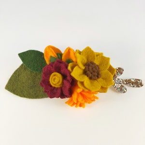 Op de afbeelding: Een haarspeld van vilt met een zonnebloem, een bordeauxrode roos en een klein oranje bloemetje. De speld heeft een groen blad en een glinsterende gouden steel.
