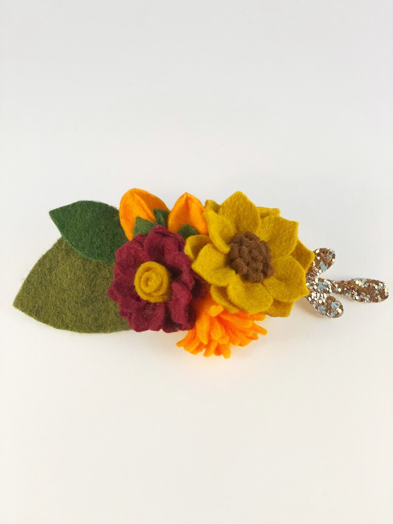 Op de afbeelding: Een haarspeld van vilt met een zonnebloem, een bordeauxrode roos en een klein oranje bloemetje. De speld heeft een groen blad en een glinsterende gouden steel.