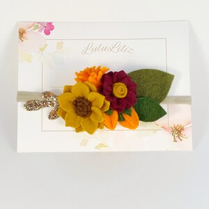 Op de afbeelding: Een haarband van vilt met een zonnebloem, een bordeauxrode bloem en een kleine oranje bloem. De haarband is beige met gouden glitter.