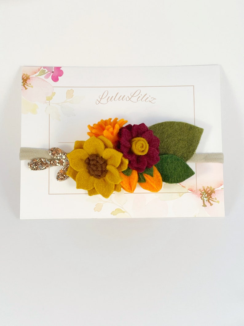 Op de afbeelding: Een haarband van vilt met een zonnebloem, een bordeauxrode bloem en een kleine oranje bloem. De haarband is beige met gouden glitter.