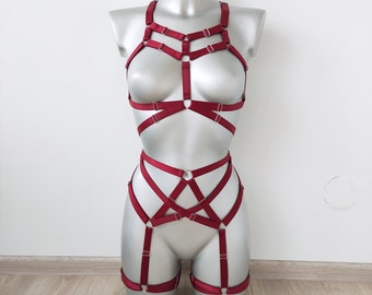 May include: A burgundy lingerie set displayed on a mannequin. The set includes a harness-style top and a matching bottom with adjustable straps and metal hardware. The design features a series of intersecting straps and rings, creating a geometric pattern.