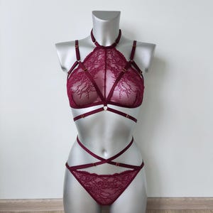 Könnte beinhalten: Ein tiefburgunderrotes Spitzen-Dessous-Set bestehend aus einem hochgeschlossenen Bralette mit Halsband, komplexen Blumenmustern und mehreren verstellbaren Trägern mit goldfarbenen Elementen. Das passende Unterteil hat überkreuzte Taillenbänder.