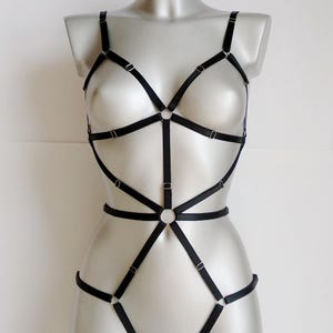 May include: Black body harness with adjustable straps and metal hardware. The harness features a geometric design with multiple connecting straps and rings. The lingerie is displayed on a mannequin against a white background.