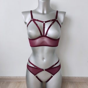 May include: A burgundy lingerie set on a mannequin. The set includes a bra with a cage design and matching high-waisted bottoms. The bra has a sheer mesh panel. The lingerie is made of a soft, sheer fabric.