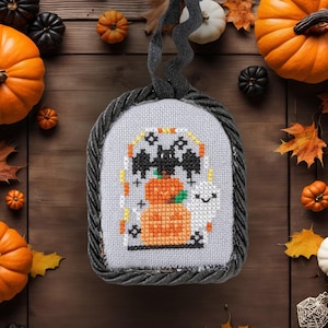 May include: A gray and black fabric ornament with a stitched design of a bat, a ghost, and pumpkins. The ornament is surrounded by fall leaves and pumpkins.