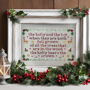 May include: A framed cross-stitch with the text "the holly and the ivy" in red thread. The design includes deer, candles, and holly sprigs. The white frame is decorated with holly and red berries, evoking a festive atmosphere.