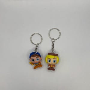 May include: Two cartoon character keychains. One keychain features a boy with blue hair, a blue hat, and a brown shirt. The other keychain features a girl with blonde hair, a brown hat, and a brown shirt.