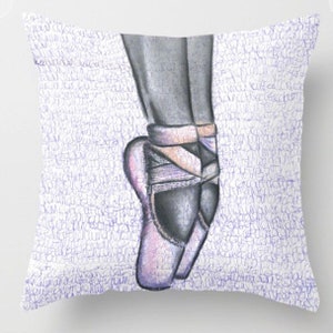 May include: Square throw pillow featuring a pencil sketch of a ballerina's feet in pointe shoes. The shoes are a soft pink with gray accents, and the background is white with a pattern of purple script.
