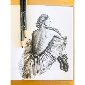 May include: A charcoal sketch of a ballerina in a tutu, viewed from the back, seated with her arms crossed. Two charcoal pencils are placed on the open sketchbook. The drawing is in shades of gray and black.