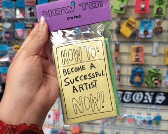 May include: A plastic bag containing a set of yellow, green, and blue cards with the text "HOW TO: BECOME A SUCCESSFUL ARTIST NOW!" printed on them. The cards are labeled "Zine Pack".