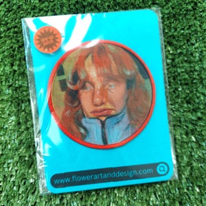May include: A red and white sew-on patch featuring a portrait of a person with red hair. The patch is on a blue background with the text "www.flowerartanddesign.com" and a magnifying glass icon.