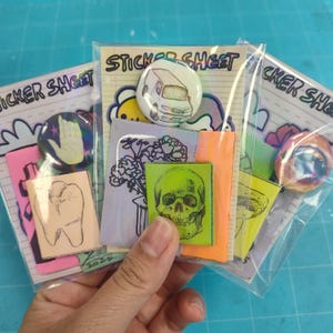 May include: A collection of sticker sheets and button pins. The sticker sheets feature various illustrations, including a tooth, flowers, and a skull. The button pins have a hand design and a van illustration. The text "STICKER SHEET" is visible.