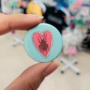May include: A blue and pink button with a heart-shaped design featuring a brown cockroach inside. The heart has a white scalloped border.