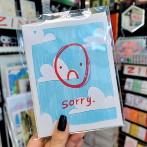 May include: A blue and white card with a red circle containing a sad face and the word "sorry" written in red.