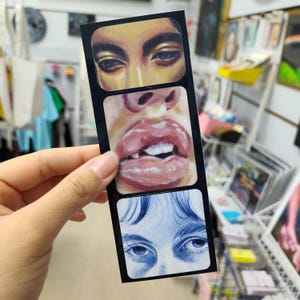 May include: A vertical rectangular sticker featuring three stacked artistic panels. The top panel shows a pair of brown eyes, the middle panel displays a close-up of glossy lips with visible teeth, and the bottom panel depicts blue-toned eyes.