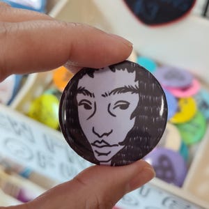 May include: A round button with a black and white illustration of a person's face. The button is held by a hand, with other colorful buttons visible in the background. The face has a light purple skin tone and black hair.