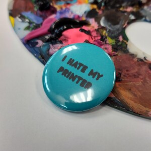 May include: A round, teal button with the phrase "I HATE MY PRINTER" in black text. The button is glossy and sits on a white surface, with a colorful artist's palette in the background.