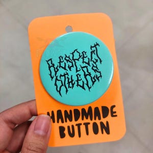 May include: A turquoise blue button with black text that reads "Respect Others" in a gothic font. The button is on a bright orange card that says "Handmade Button".