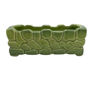 May include: A rectangular, green ceramic planter with a textured, stone-like exterior. The planter has a slightly scalloped top edge and sits on four small feet. The color is a vibrant, light green.