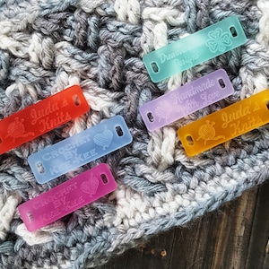 May include: A collection of colorful acrylic tags for handmade knit and crochet items. The tags are in various colors, including red, blue, purple, and yellow, with engraved text and symbols like yarn and hearts. The tags are placed on a textured, gray and white knitted fabric.