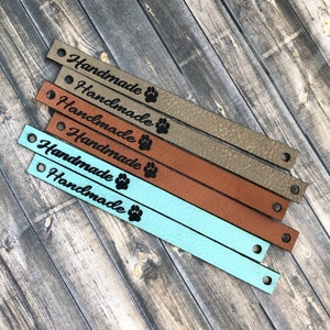 May include: A collection of leather straps in various colors, including tan, brown, and teal. Each strap is engraved with the word "Handmade" and a paw print. The straps have holes at each end.