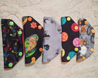 May include: Five fabric squares with different patterns and colors. The squares have snap buttons on the corners. The patterns include musical notes, a car, a building, smiley faces, and a skull.