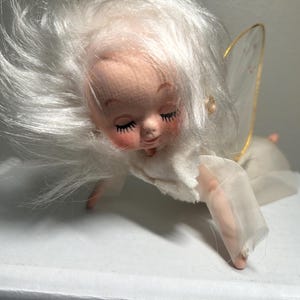 May include: A vintage angel doll with a soft, fabric face, closed eyes, and rosy cheeks. The doll has white hair, a white dress, and sheer, gold-trimmed wings. The doll is lying down, with one arm extended.