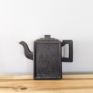 May include: Black teapot with a square body and a raised design of a person and a bird. The teapot has a spout and a handle.