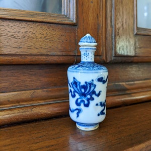 May include: A small, white porcelain bottle with a blue and white floral design. The bottle has a lid with a small knob on top. The bottle is sitting on a wooden surface.