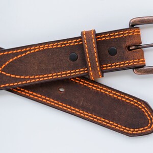 May include: A dark brown leather belt featuring vibrant orange contrast stitching in a decorative wave pattern. The belt includes a rectangular, weathered copper-toned metal buckle and two black rivets securing the leather loop near the buckle.