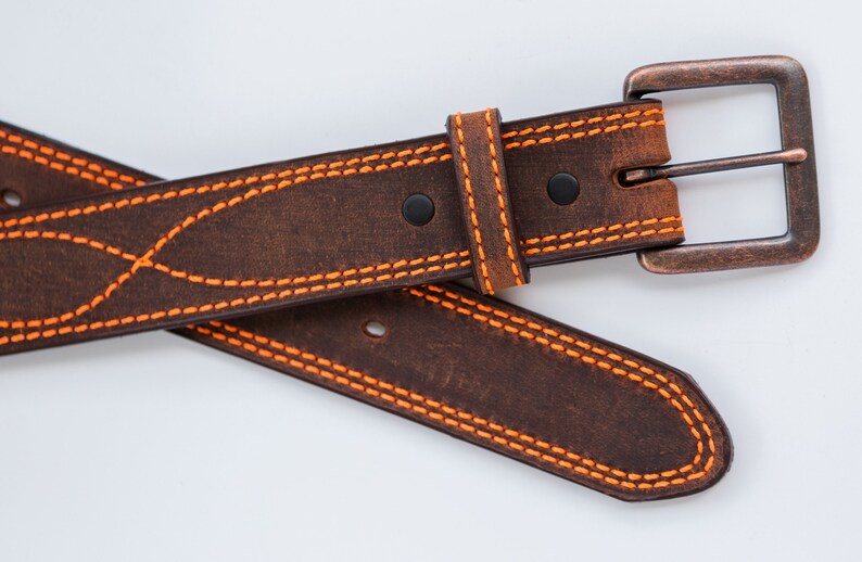 May include: A dark brown leather belt featuring vibrant orange contrast stitching in a decorative wave pattern. The belt includes a rectangular, weathered copper-toned metal buckle and two black rivets securing the leather loop near the buckle.