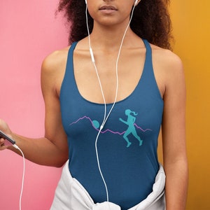 May include: A woman wearing a blue tank top with a graphic of a person running on a mountain path. The graphic is in teal and pink. The woman is wearing white headphones and holding a black smartphone.