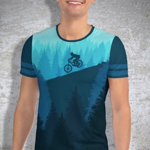 May include: A blue t-shirt featuring a mountain biking scene. The design shows a cyclist on a bike against a backdrop of a forest and sky in shades of blue. The shirt has a crew neck and striped sleeves.
