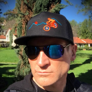 Puede incluir: Una gorra de camionero negra con un diseño bordado en azul y naranja de una persona montando una bicicleta de montaña sobre una cordillera. La gorra tiene una visera negra y una parte trasera de malla.