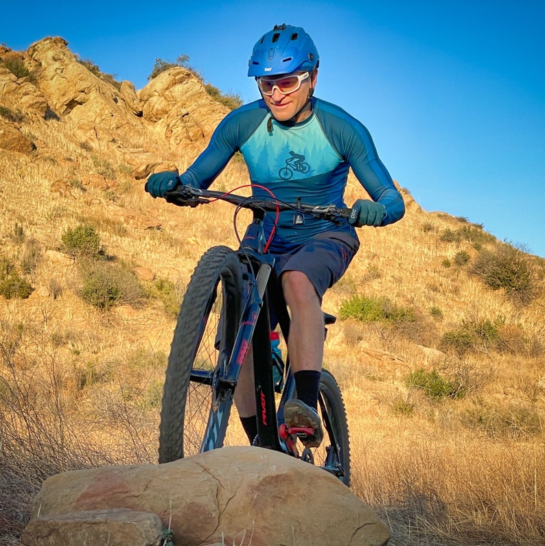 May include: A person wearing a blue long-sleeved shirt with a bicycle graphic rides a black mountain bike with a blue helmet on a dirt path.