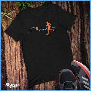 May include: A black t-shirt featuring a graphic of a person running in front of a mountain range with a sun setting behind the mountains. The graphic is in orange and blue.