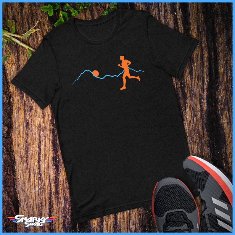 May include: A black t-shirt featuring a graphic of a person running in front of a mountain range with a sun setting behind the mountains. The graphic is in orange and blue.