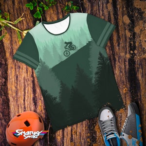May include: A green t-shirt with a mountain biking design. The shirt features a silhouette of a cyclist on a mountain, with a forest backdrop. The shirt has a white collar and green sleeves with two stripes. An orange helmet and sneakers are also in the image.