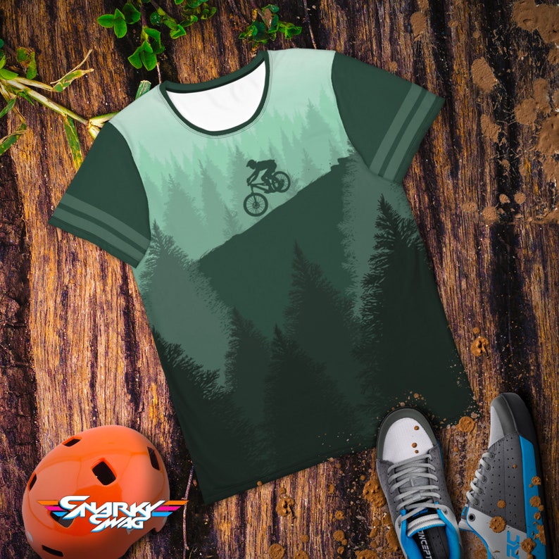 May include: A green t-shirt with a mountain biking design. The shirt features a silhouette of a cyclist on a mountain, with a forest backdrop. The shirt has a white collar and green sleeves with two stripes. An orange helmet and sneakers are also in the image.