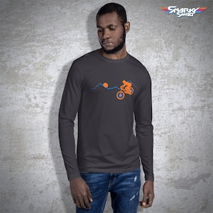 May include: A dark gray long-sleeve t-shirt with an orange silhouette of a person riding a bicycle over a mountain range with a setting sun. The text "SNARKY SWAG" is in the upper right corner of the image.