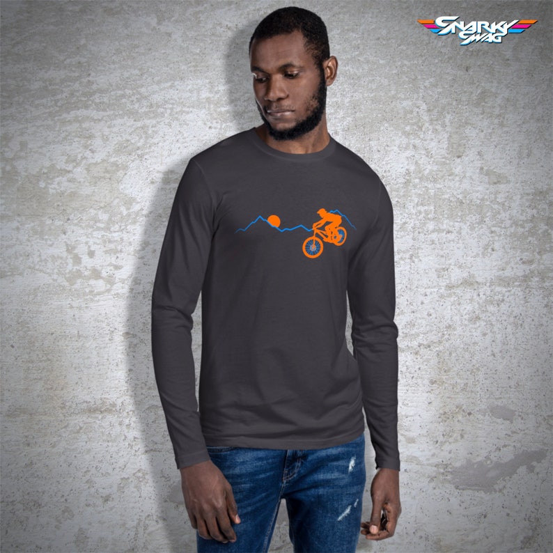 May include: A dark gray long-sleeve t-shirt with an orange silhouette of a person riding a bicycle over a mountain range with a setting sun. The text "SNARKY SWAG" is in the upper right corner of the image.