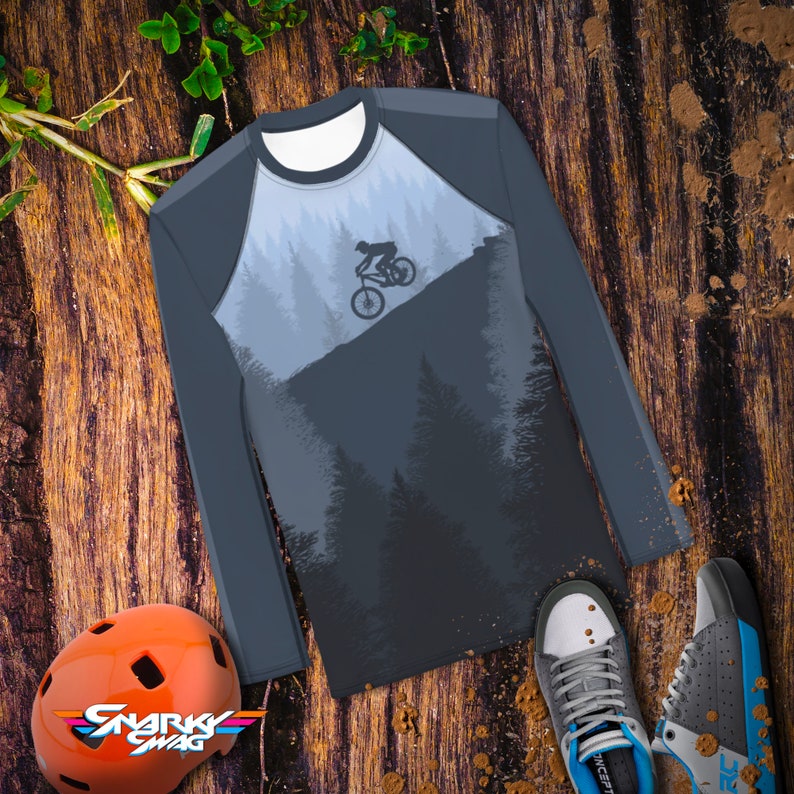 May include: A long-sleeved shirt with a dark blue and gray color scheme. The shirt features a graphic of a person riding a mountain bike through a forest. The text "Snarky Swag" is printed on a helmet.