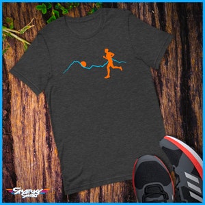 May include: A dark heather grey t-shirt featuring a graphic of a person running in front of a mountain range with a sun setting behind the mountains. The graphic is in orange and blue.