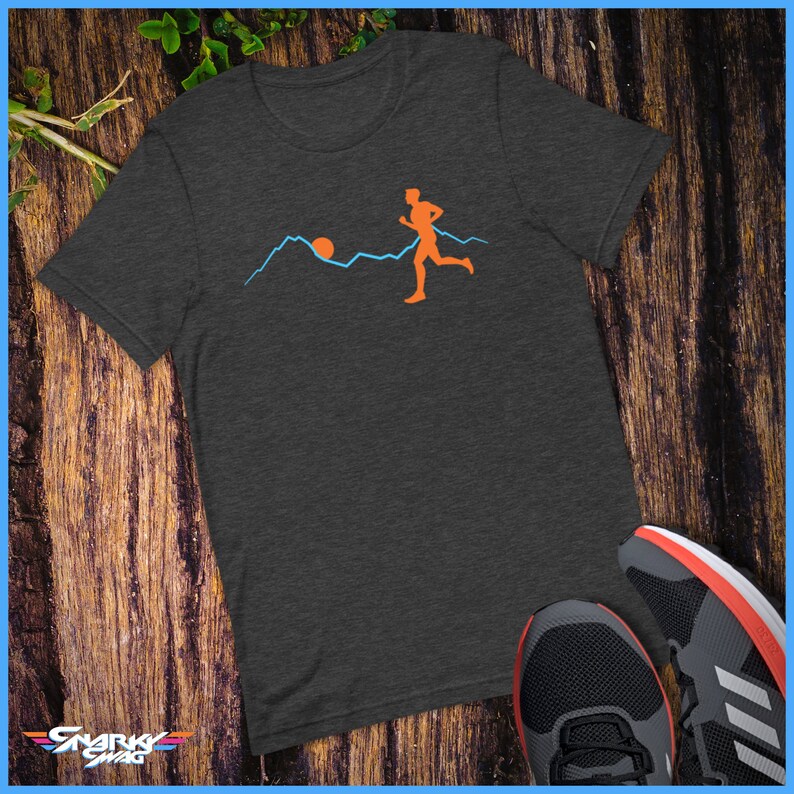 May include: A dark heather grey t-shirt featuring a graphic of a person running in front of a mountain range with a sun setting behind the mountains. The graphic is in orange and blue.