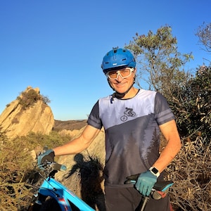 May include: A blue mountain bike with a black seat and blue frame. The person is wearing a blue helmet, white glasses, and a blue and gray shirt with a mountain biking graphic. The gloves are blue and the watch is silver.