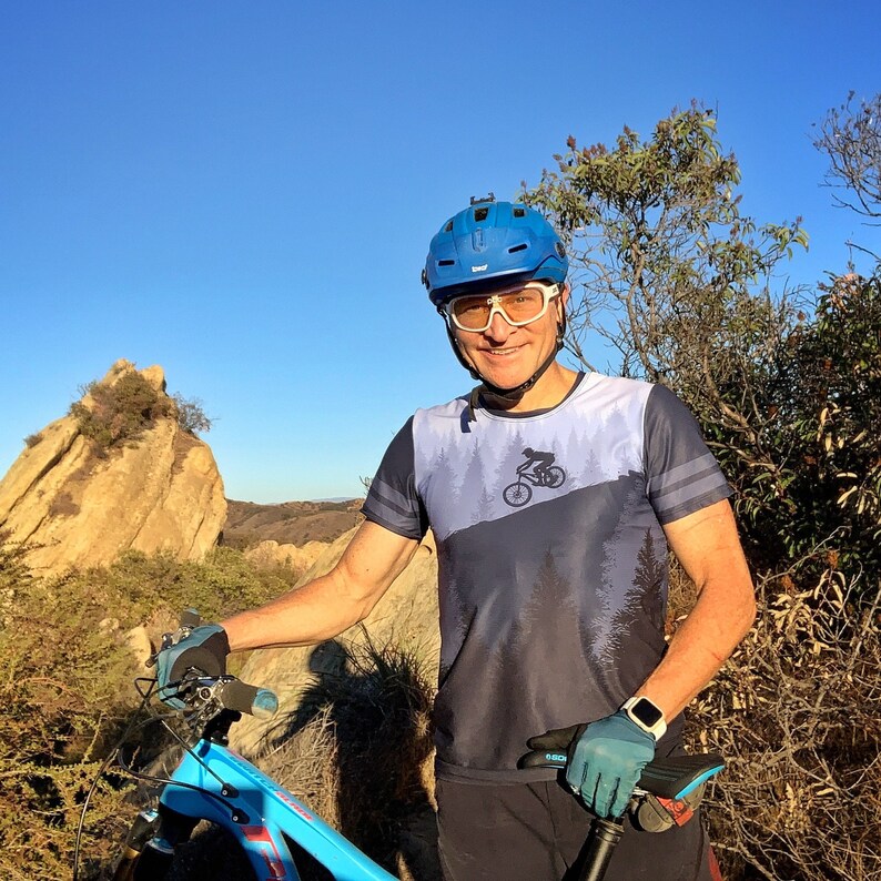May include: A blue mountain bike with a black seat and blue frame. The person is wearing a blue helmet, white glasses, and a blue and gray shirt with a mountain biking graphic. The gloves are blue and the watch is silver.