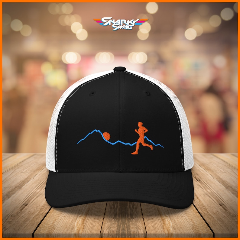Puede incluir: Gorra de camionero negra y blanca con un dise&ntilde;o bordado en negro de una persona corriendo por una cordillera con el sol saliendo detr&aacute;s de ella. El dise&ntilde;o es en naranja y azul.